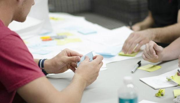 Zwei Personen sitzen an einem Tisch und basteln mit Papier. Kleine Knöllchen liegen herum, wahrscheinlich von zerknüllten Post Its.