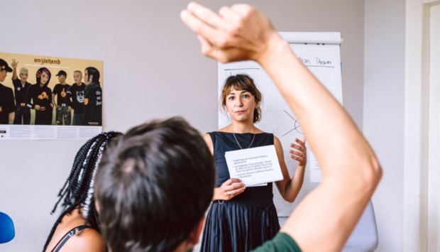 Eine Frau im schwarzen Kleid steht vor einem Flipchart und hat während ihrem Vortrag Moderationskarten in der Hand. Ein Teilnehmer meldet sich.