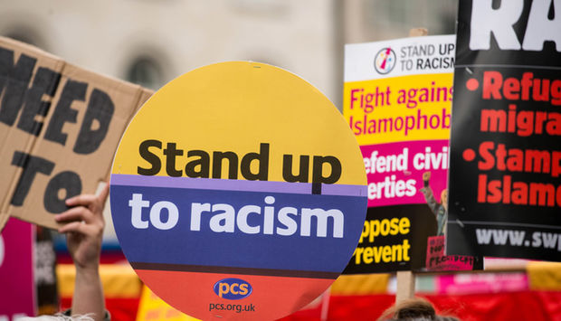 Ein Foto von einer Demonstration, viele Transparente, im Vordergrund ein Schild mit den Worten: Stand up to racism