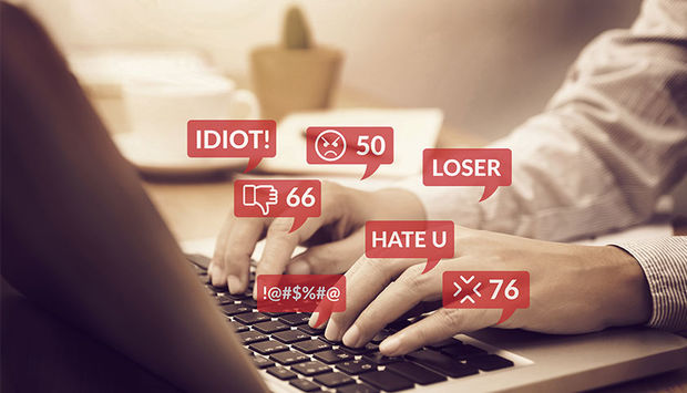 Ein Foto mit hellen und warmen Farben von einem Menschen, der am Laptop arbeitet. Über der Tatstatur sind rote Sprechblasen, in denen mit weißen Buchstaben zum Beispiel Hate U, Loser, Idiot steht. Es ist ein Symbolbild für Hassrede im Internet.