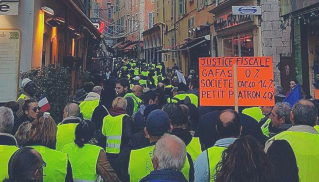 Ein Foto von einer Demonstration der Gelbwesten-Bewegung in Frankreich mit vielen Menschen mit gelben Warnwesten in einer engen Gasse udn einem Organgen Schid.