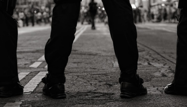 Ein Bild von einem breitbeinig stehenden Mann von hintern knieabwärts. Man sieht durch die BEine die Straße und es könnte eine Demonstration sein. Sinnbild für extreme Rechte und Männlichkeit.