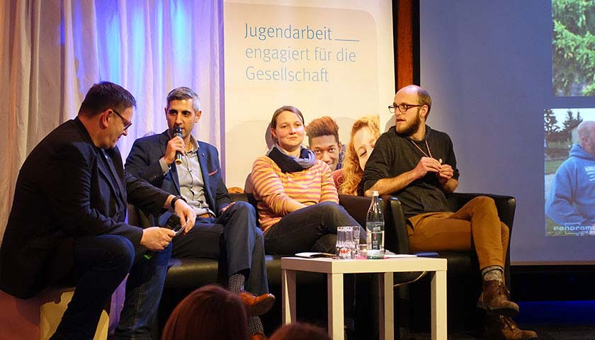 Abgebildet sind 4 Personen auf einem Podium, ein Mann hält ein Mikrofon.