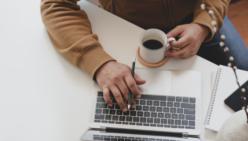 Eine Person sitz vor einem Laptop und hält eine Kaffeetasse in der Hand.